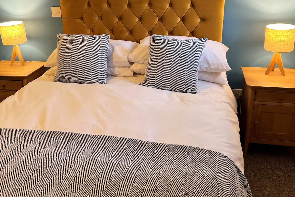 Una cama bien tendida con un cabecero capitonado, dos lámparas de noche y una colcha estampada.