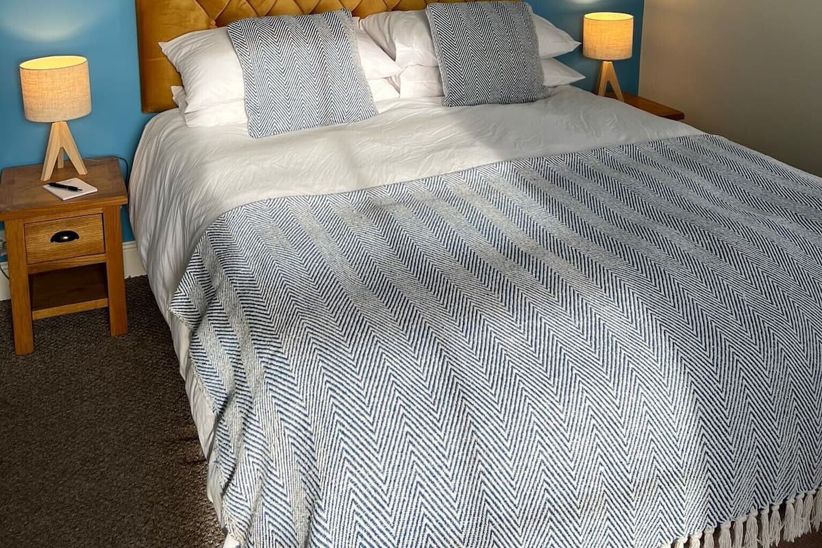 Un dormitorio con pared azul, cabecera de madera, cama con ropa de cama blanca y azul, y dos lámparas de noche.