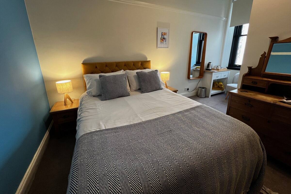 Habitación de hotel ordenada con una cama grande, lámparas de noche, un aparador, un espejo y un cuadro en la pared.
