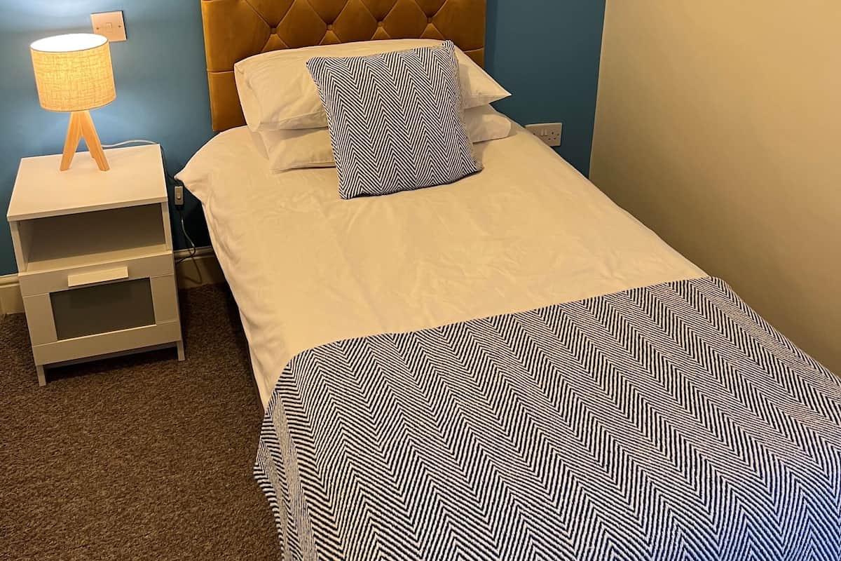 Una cama impecable con un edredón estampado, un cabecero capitonado y una lámpara de noche sobre una mesita.