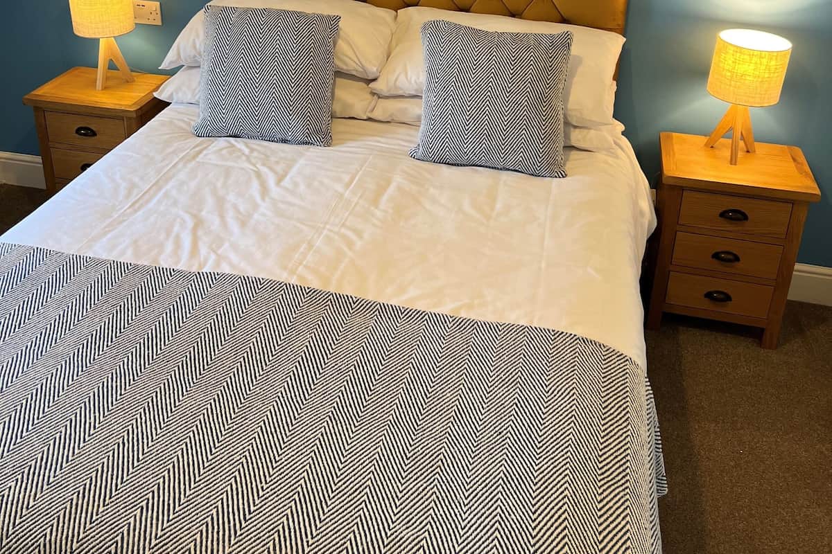 Una cama bien hecha con una colcha estampada y dos almohadas a juego, flanqueada por dos mesitas de noche con lámparas, en una habitación con paredes azules y techo inclinado.