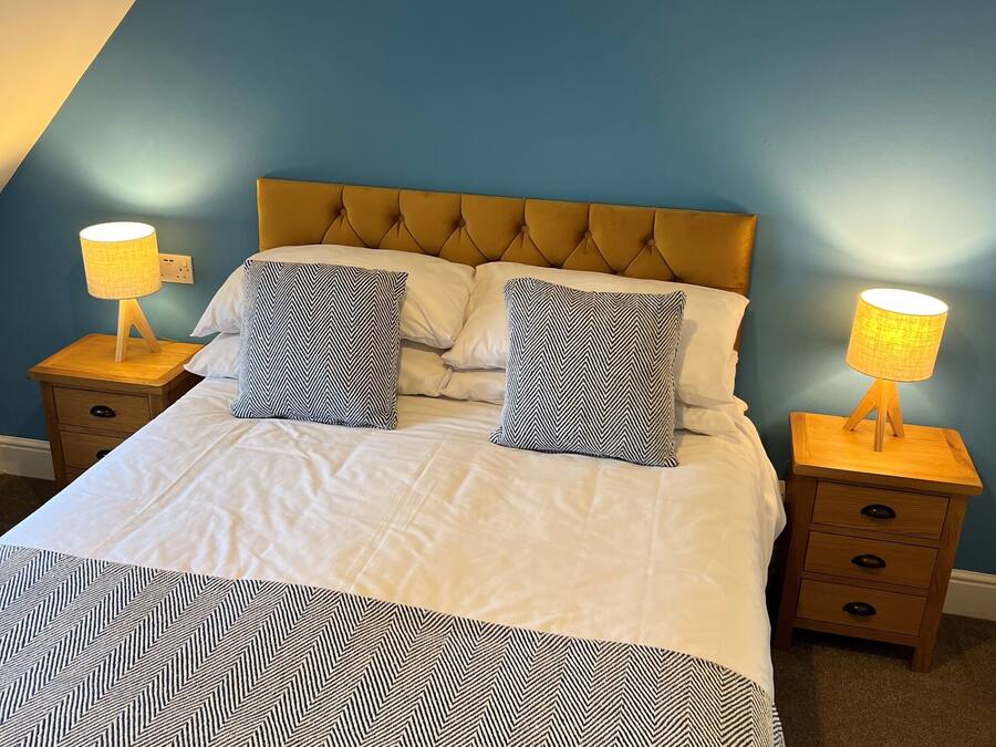 A neatly made bed with a patterned bedspread and two matching pillows, flanked by two bedside tables with lamps, in a room with blue walls and a sloped ceiling.