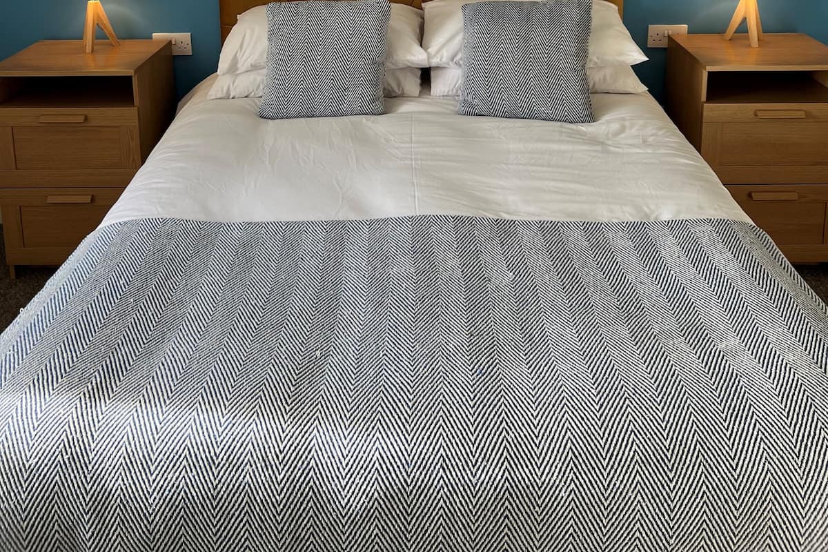 Una cama bien hecha con una colcha estampada, dos almohadas a juego y un cabecero capitonado. Hay dos mesitas de noche con lámparas, y las paredes están pintadas de azul.