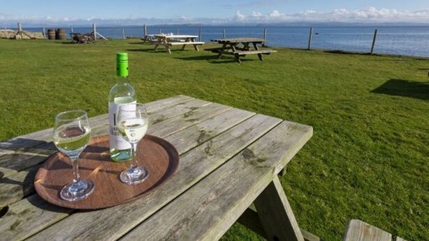 Une table de pique-nique avec une bouteille de vin blanc et deux verres, le tout posé sur un plateau en bois, est installée sur une pelouse, offrant une vue sur la mer et les montagnes en arrière-plan.