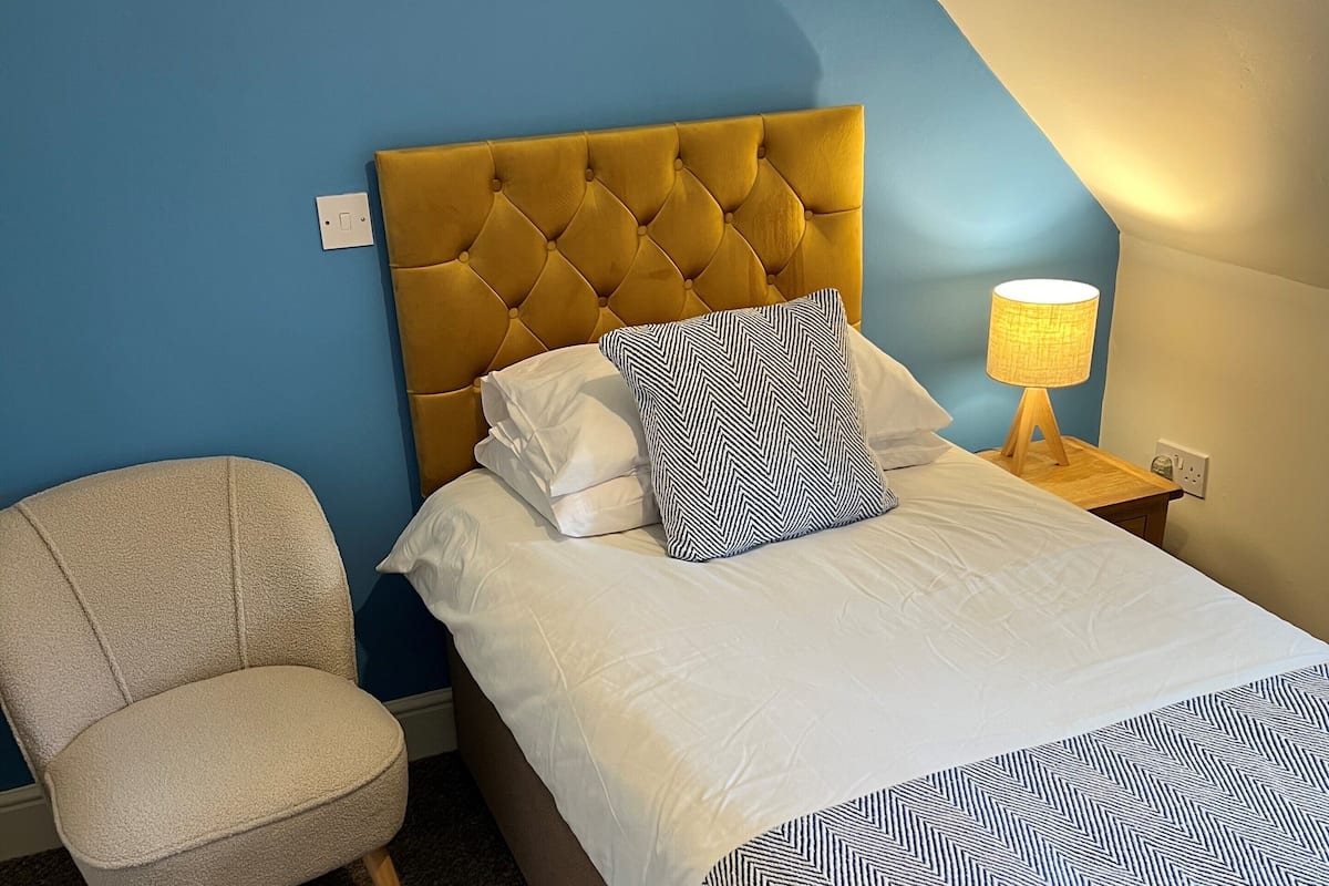 Un dormitorio con cabecera capitonada, cama con ropa de cama blanca y almohadas estampadas, una mesita de noche con lámpara y una silla.