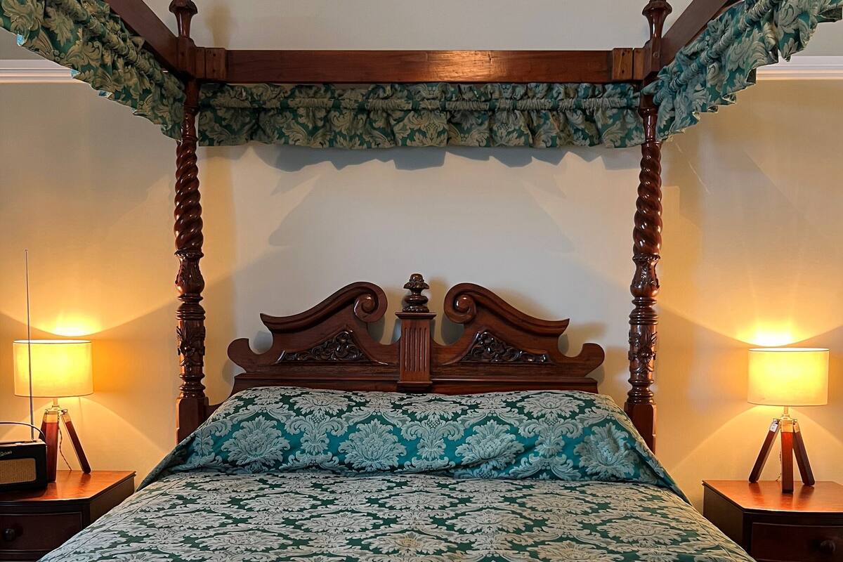 Una cama con dosel, con un edredón estampado con motivos florales y almohadas a juego, flanqueada por dos mesitas de noche con lámparas.