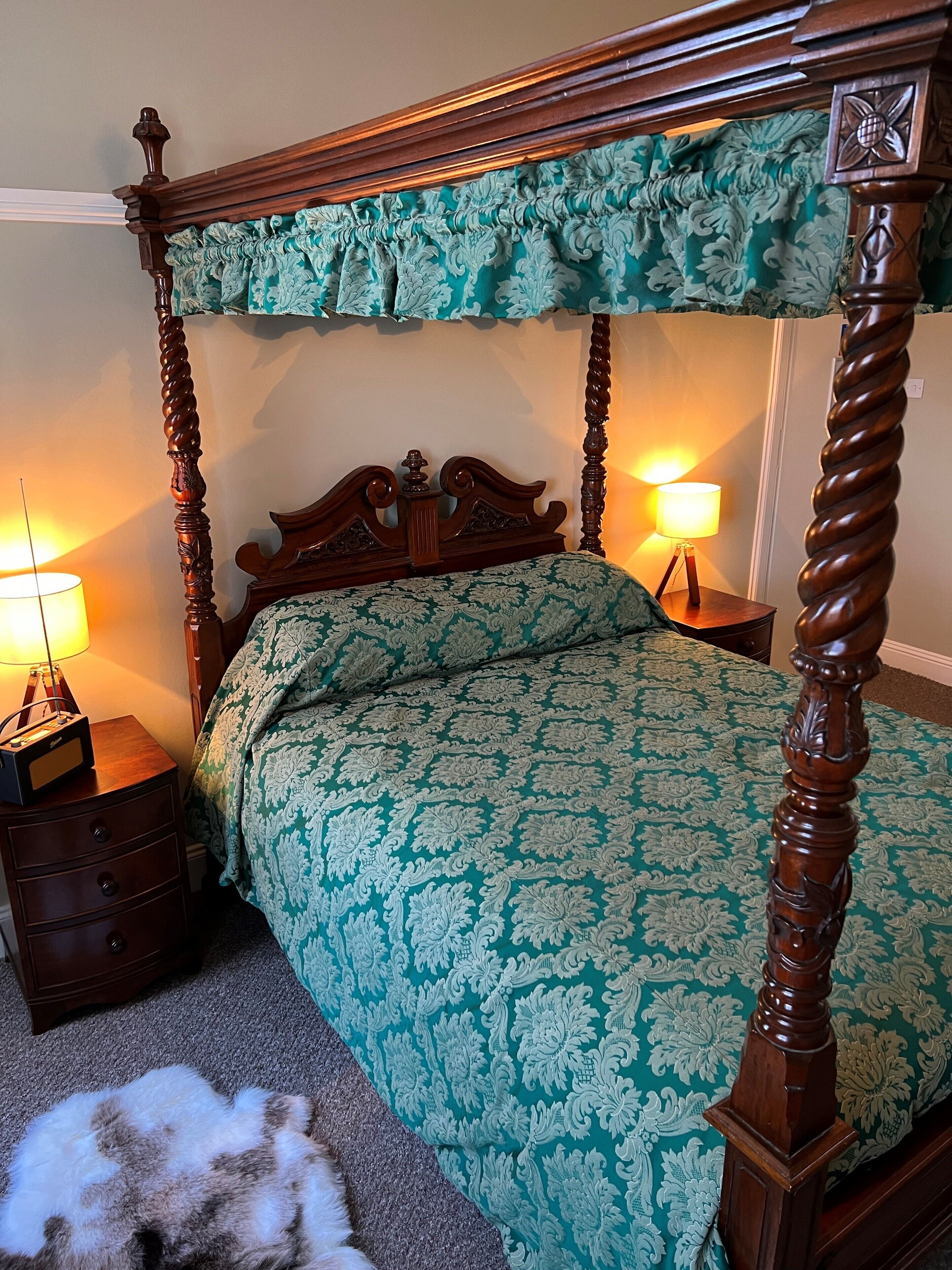 Un lit à baldaquin avec une couvre-lit à motifs verts, des tables de chevet en bois avec des lampes, et un chien blanc et duveteux allongé sur le sol.