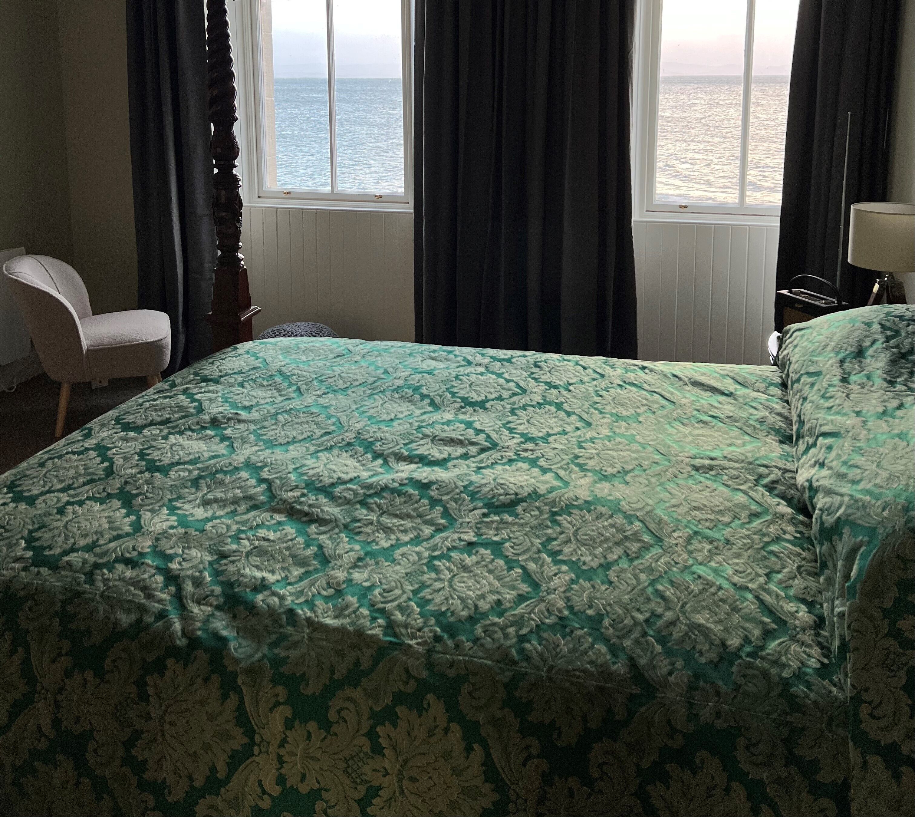 A room with a bed, a chair, and a view of the sea through the window.