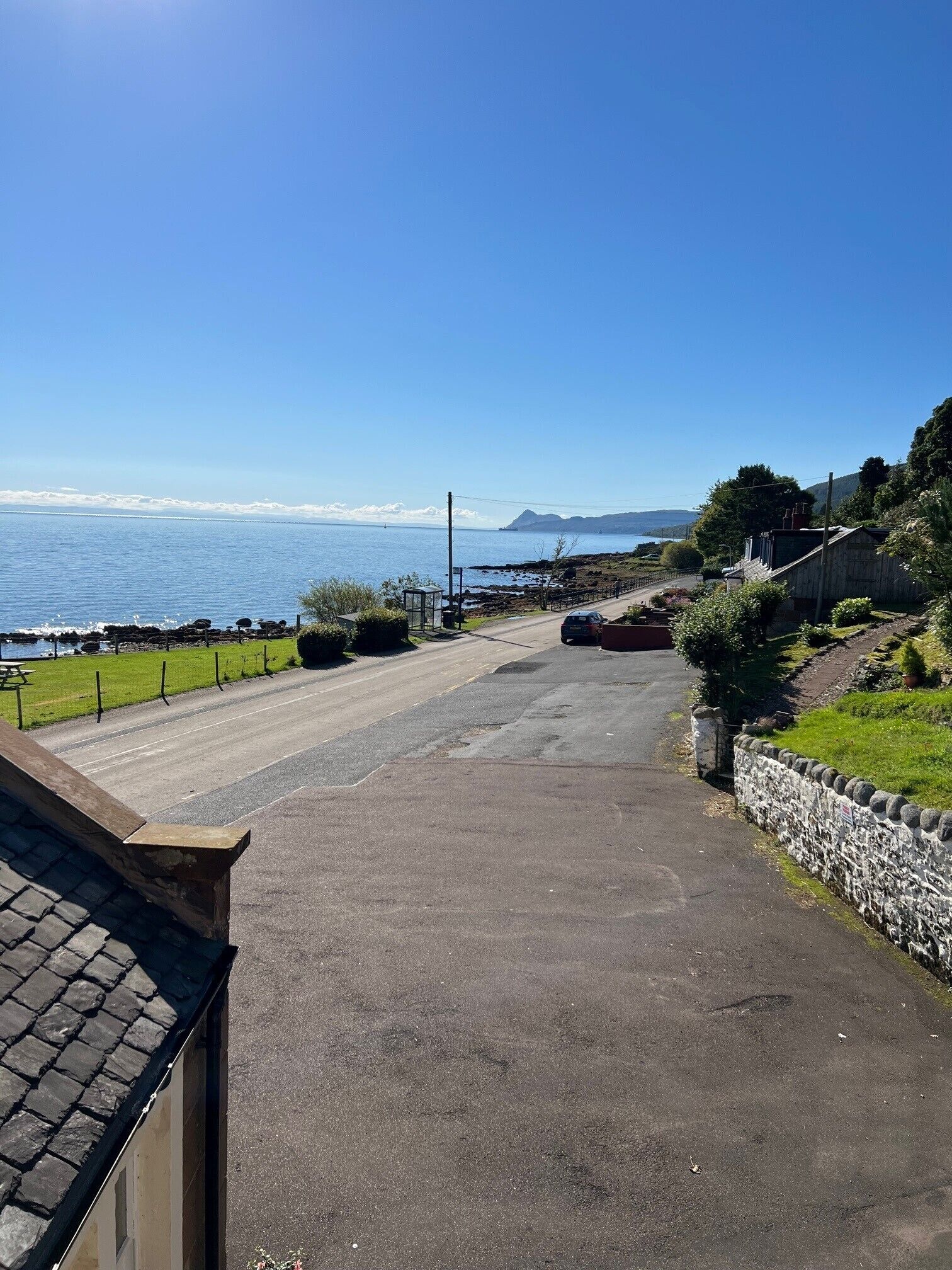 A coastal road with a stone wall and greenery on one side, leading to a body of water with a distant mountain.