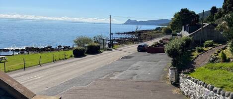 A coastal road with a stone wall and greenery on one side, leading to a body of water with a distant mountain.