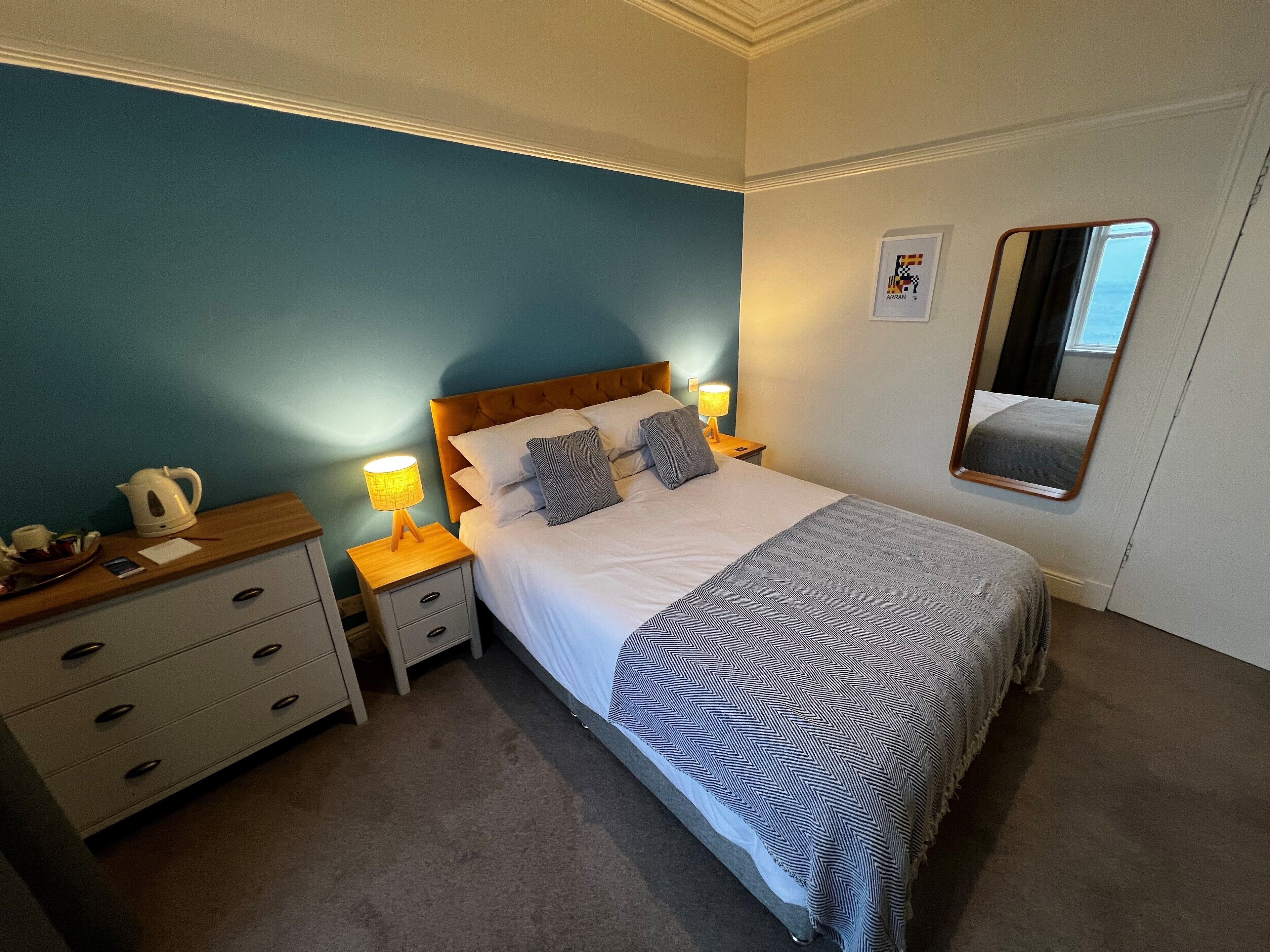 A neatly arranged bedroom with a bed, bedside tables, a nightstand, a mirror, and a wall-mounted picture.