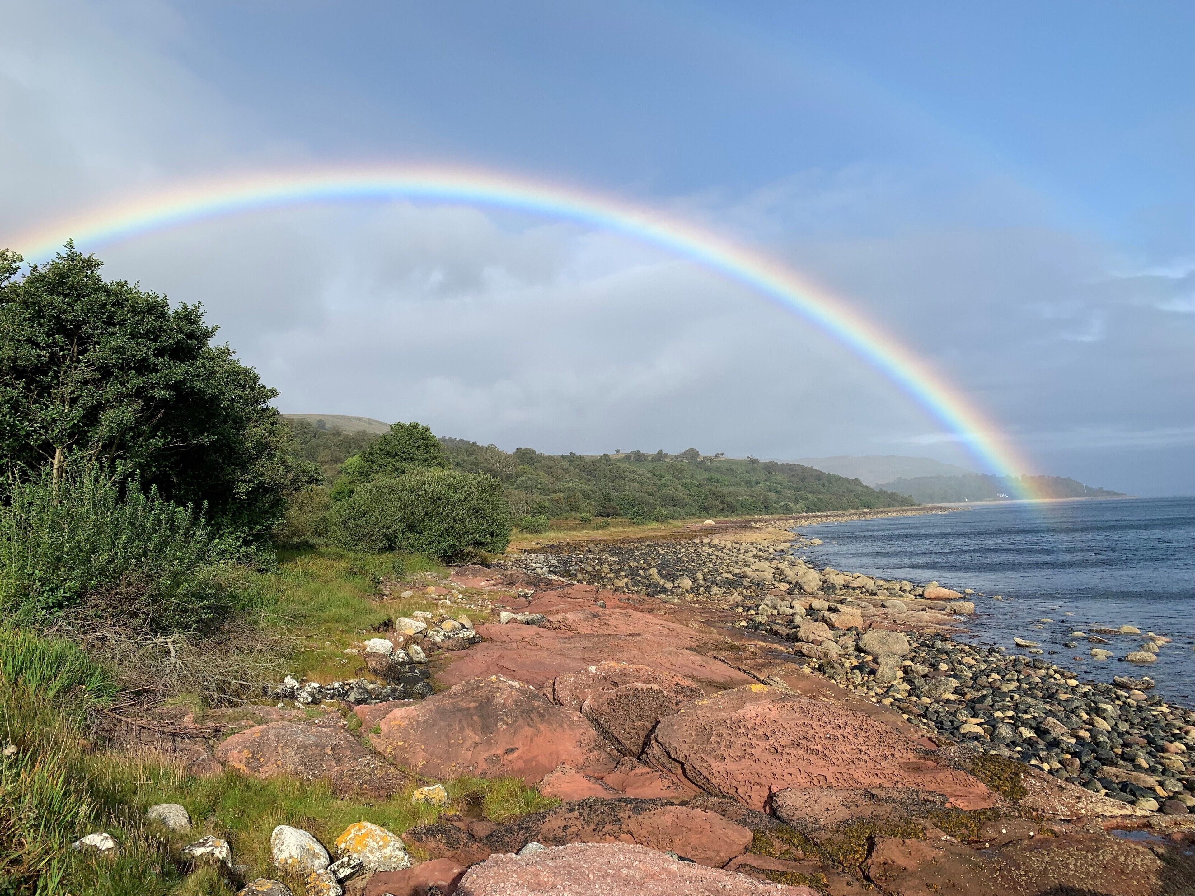A coastal landscape with a rainbow over a rocky shoreline and a forested hill in the background.
