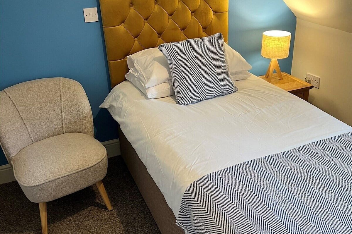 Un dormitorio con cabecera capitonada, cama con ropa de cama blanca, manta estampada, una silla y una lámpara de noche.