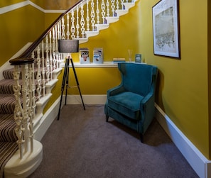 Una escalera con barandillas blancas ornamentadas, un sillón azul turquesa y un trípode con una cámara en el suelo.