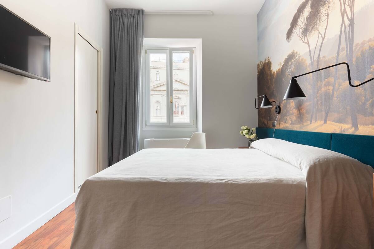 A bedroom with a bed, a desk, a chair, a TV, and a window with curtains.