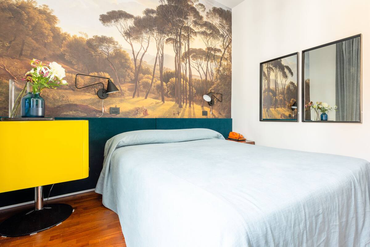 A bedroom with a bed, a desk with a lamp, a mirror, and a wall mural.