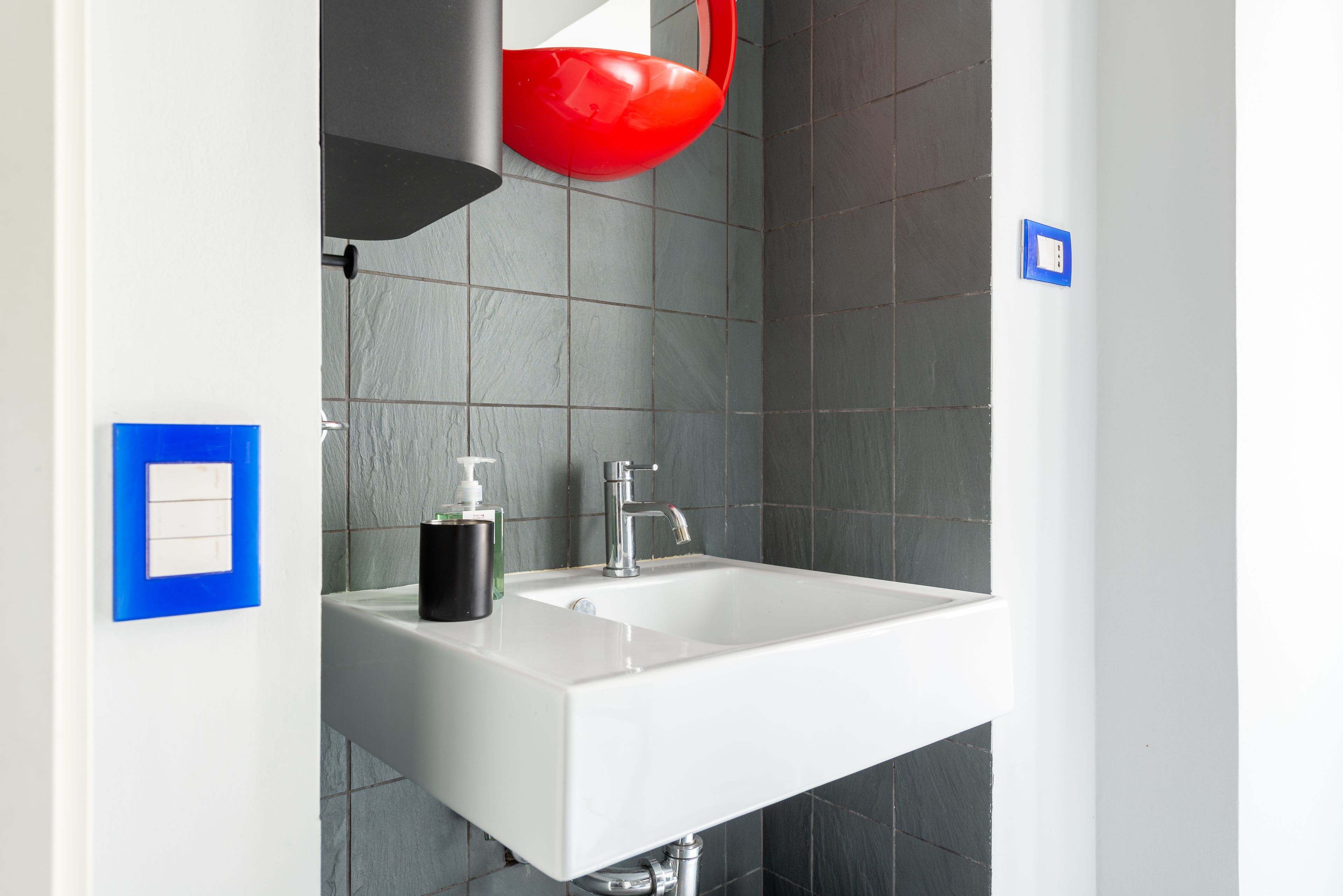 Une salle de bain moderne avec un lavabo blanc, un seau rouge, un distributeur de savon noir et un interrupteur bleu sur le mur.