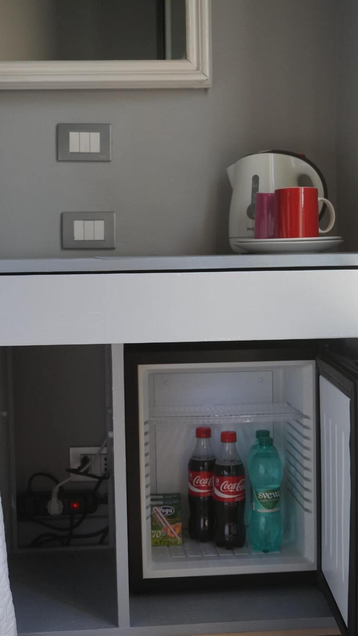 A room with a white-framed mirror, a red mug, and a refrigerator containing beverages and a box of cereal.