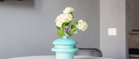 Ein runder weißer Tisch mit einer türkisfarbenen Vase mit weißen Blumen, einer weißen Pendelleuchte und einem Flachbildfernseher an der Wand.