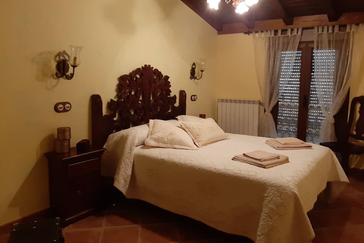 A bedroom with a wooden headboard, a bed with white bedding, a nightstand with a lamp, and a window with curtains.