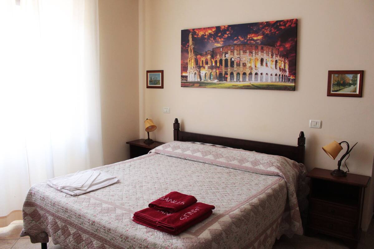 A hotel room with a bed, a nightstand, a lamp, and a painting of a historical building on the wall.