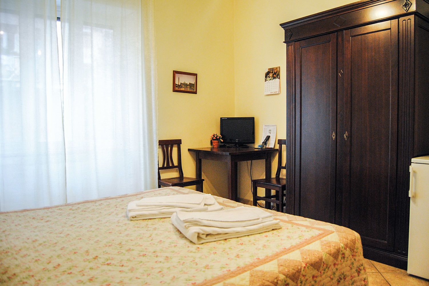 A hotel room with a bed, a desk with a computer, a wardrobe, and a small refrigerator.
