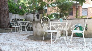 An outdoor seating area with white metal chairs and tables, surrounded by a stone wall and greenery. - Ianus Guest House (Rome)