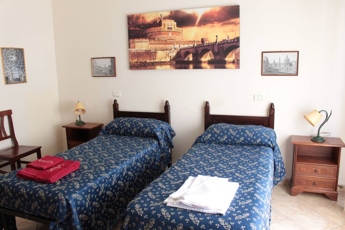 A hotel room with two beds, a nightstand, a chair, and a painting of a bridge.