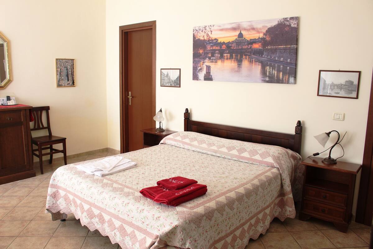 A neatly made bed with a floral bedspread and red towels, a wooden nightstand with a lamp, a chair, and a desk with a mirror and a small table.