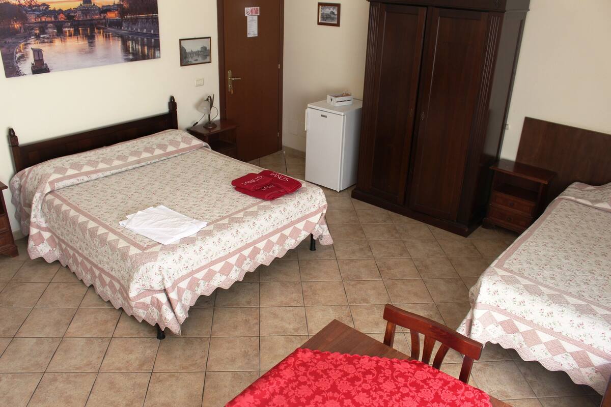 A hotel room with two beds, a small refrigerator, a wooden wardrobe, and a desk with a chair.