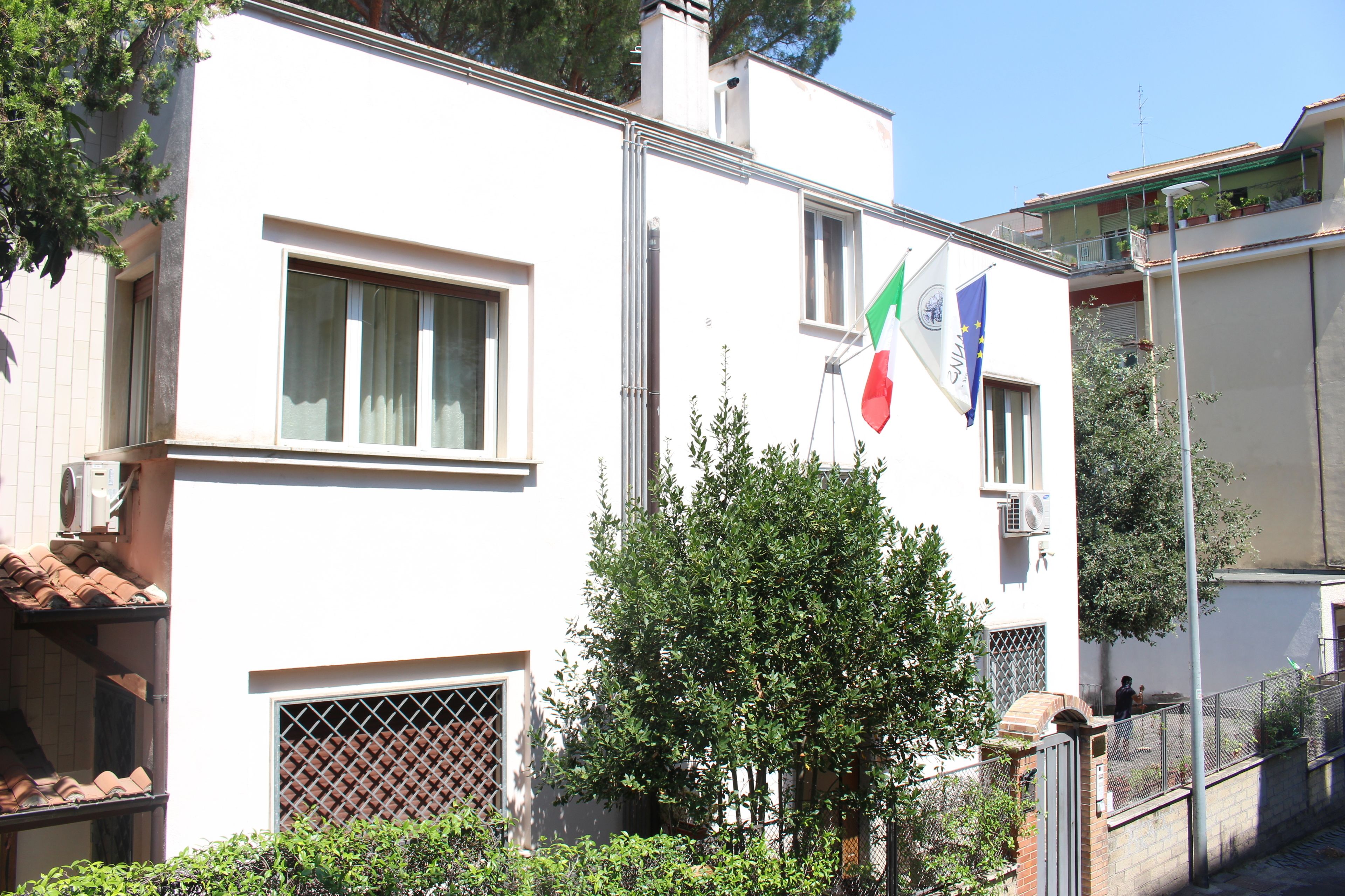 Un bâtiment blanc avec un balcon, un drapeau et un arbre devant.