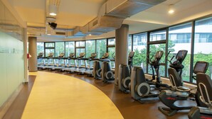 A gym with treadmills and elliptical machines arranged in rows, large windows with a view of greenery, and a curved wooden floor. - Saigon City View Apartment & Pool (Ho Chi Minh City)