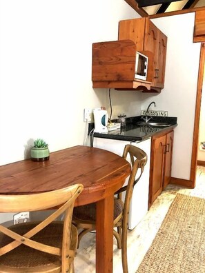 A compact kitchen with wooden cabinets, a microwave, a sink, and a dining area with a table and chairs. - Cornerstone B&B (Moqhaka)