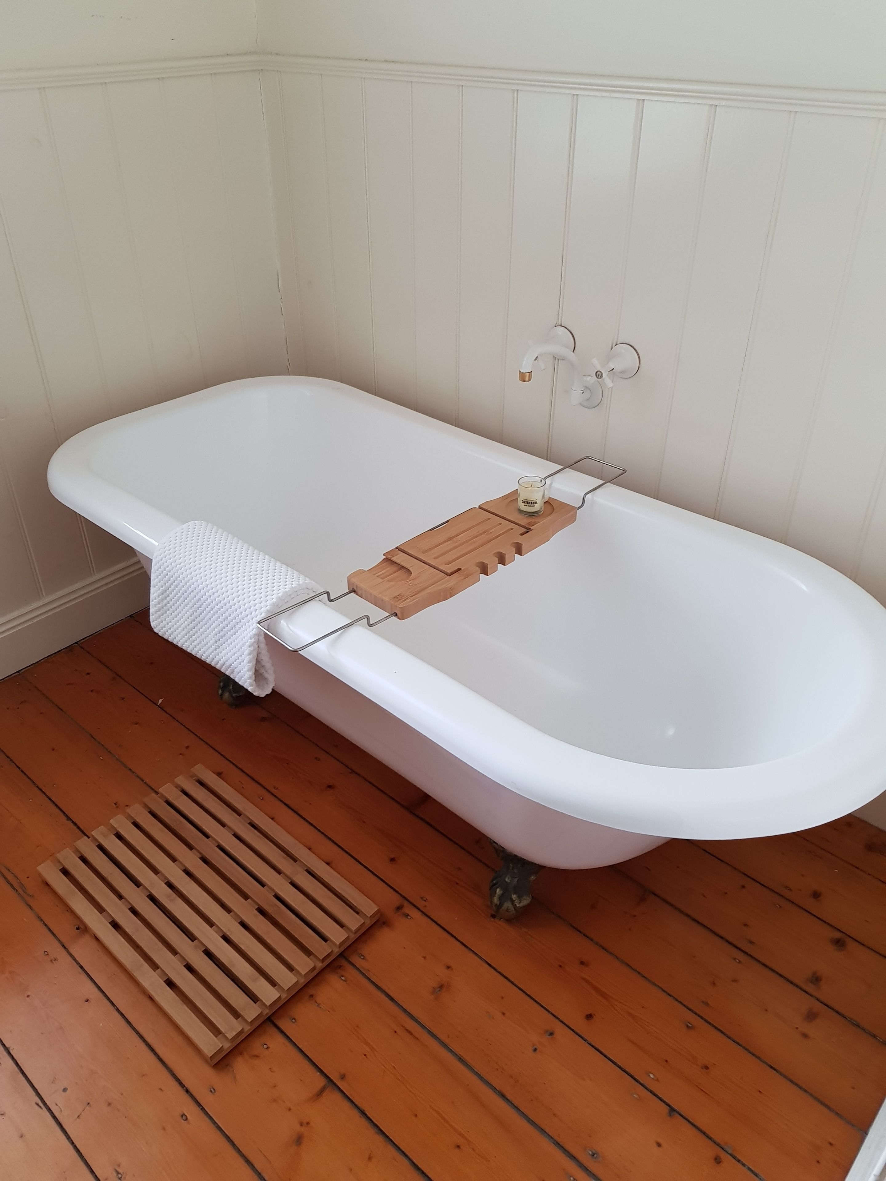 A white freestanding bathtub with a wooden tray and candle holder, set against a white paneled wall, on a wooden floor.