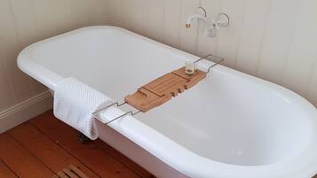 A white freestanding bathtub with a wooden tray and candle holder, set against a white paneled wall, on a wooden floor.