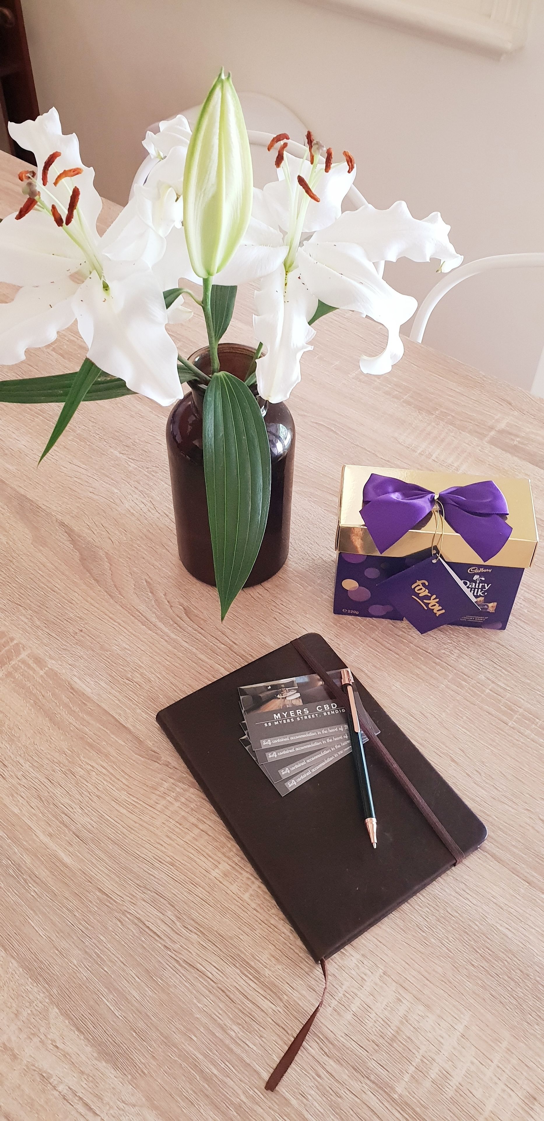 Una mesa de madera con un jarrón marrón que contiene flores blancas, una libreta negra, un bolígrafo, una pequeña caja de regalo y una barra de chocolate.