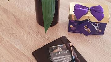 Una mesa de madera con un jarrón marrón que contiene flores blancas, una libreta negra, un bolígrafo, una pequeña caja de regalo y una barra de chocolate.