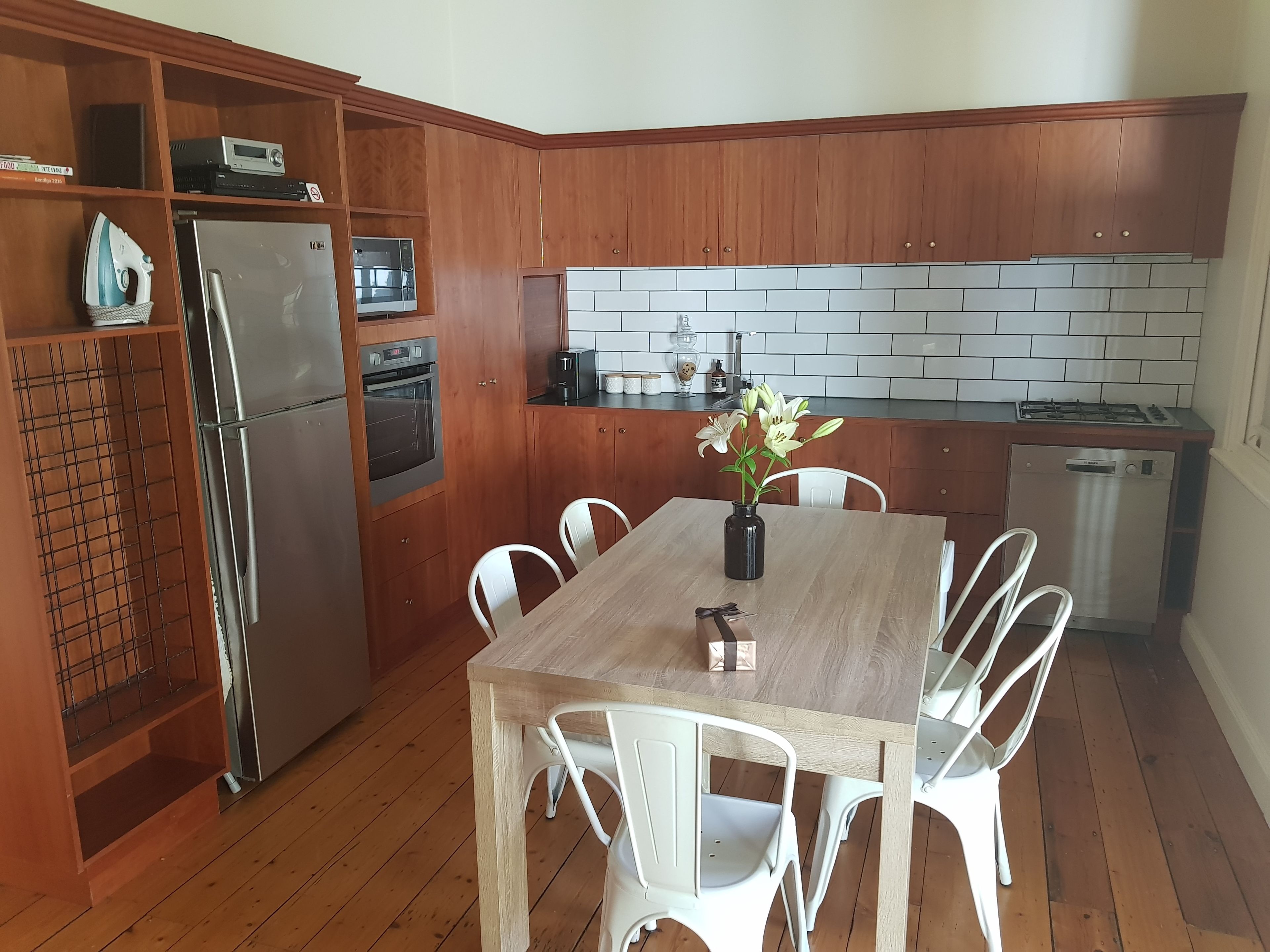 Una cocina compacta con gabinetes de madera, un refrigerador de acero inoxidable, un horno, un microondas, una estufa a gas y un lavavajillas. Hay una mesa de comedor con sillas blancas y un florero con flores.