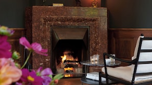 A fireplace with a lit fire, a chair, a table with a candle, and a framed picture on the wall. - Signau House & Garden (Zürich)