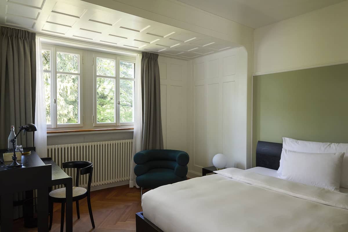A hotel room with a bed, a desk with a chair, a window with curtains, and a radiator.