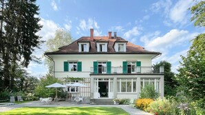 A two-story house with a balcony, a garden with outdoor furniture, and a clear sky. - Signau House & Garden (Zürich)