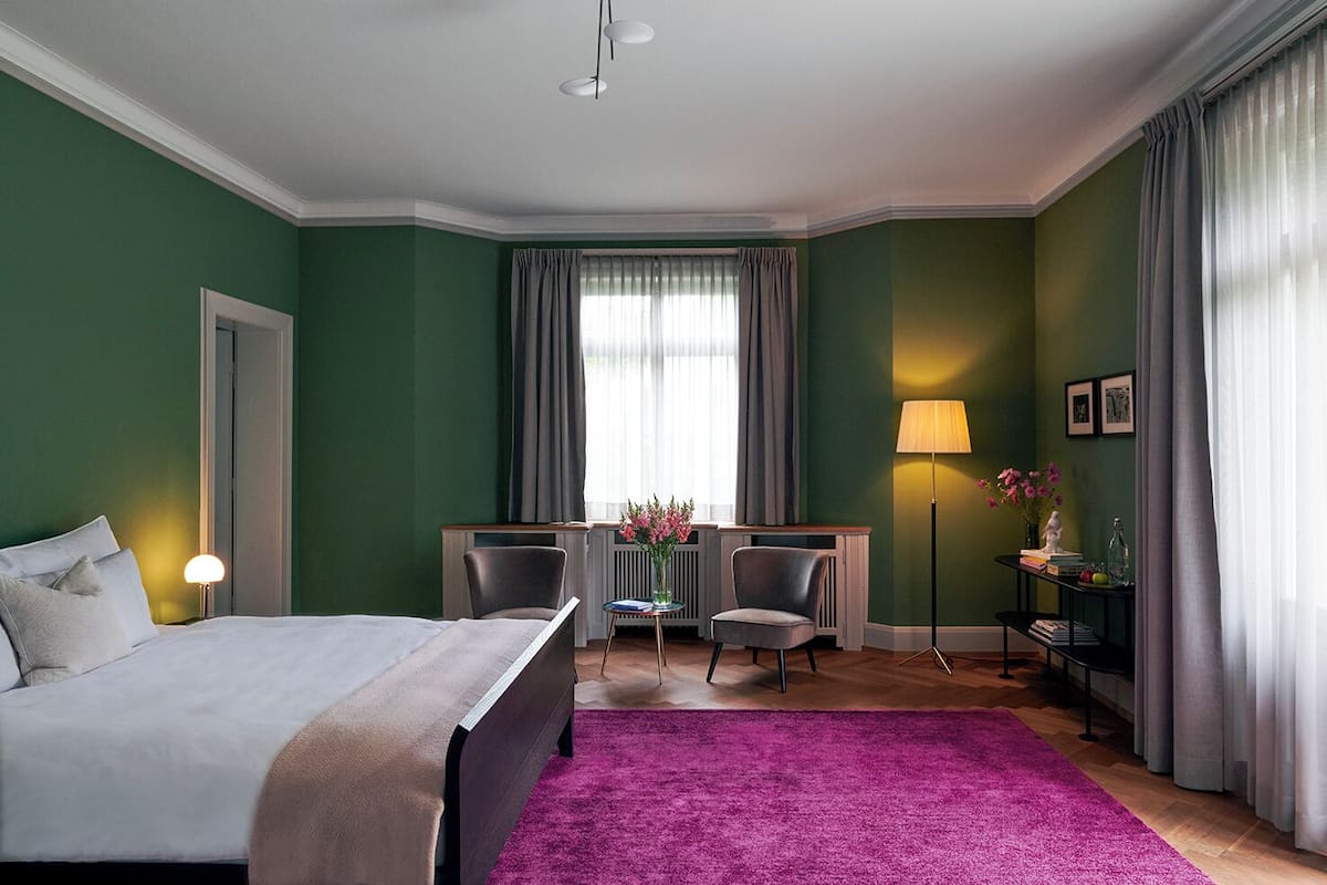 A hotel room with a bed, a desk, two chairs, a small table with flowers, a lamp, and a purple rug.