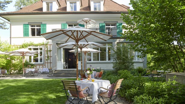 A patio with a table and chairs, surrounded by greenery and a house in the background. - Signau House & Garden (Zürich)