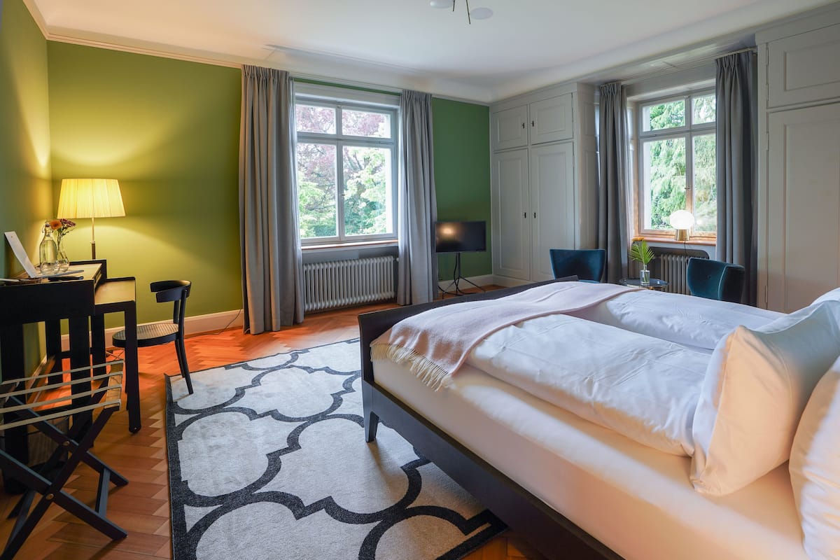 A hotel room with a large bed, a desk with a lamp, a chair, and a view of greenery outside.