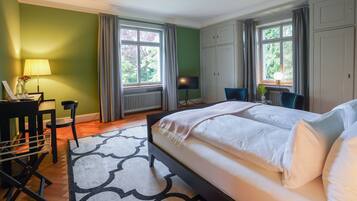 A hotel room with a large bed, a desk with a lamp, a chair, and a view of greenery outside.
