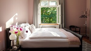 A bedroom with a bed, a nightstand with a lamp, a chair, a coat rack, and a window with a view of trees. - Signau House & Garden (Zürich)