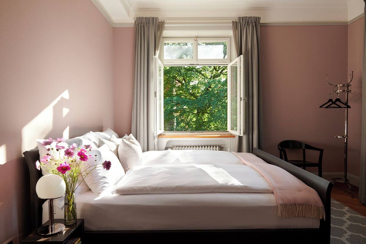 A bedroom with a bed, a nightstand with a lamp, a chair, a coat rack, and a window with a view of trees.