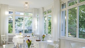 A dining area with white tables and chairs, large windows with curtains, and a checkered floor. - Signau House & Garden (Zürich)