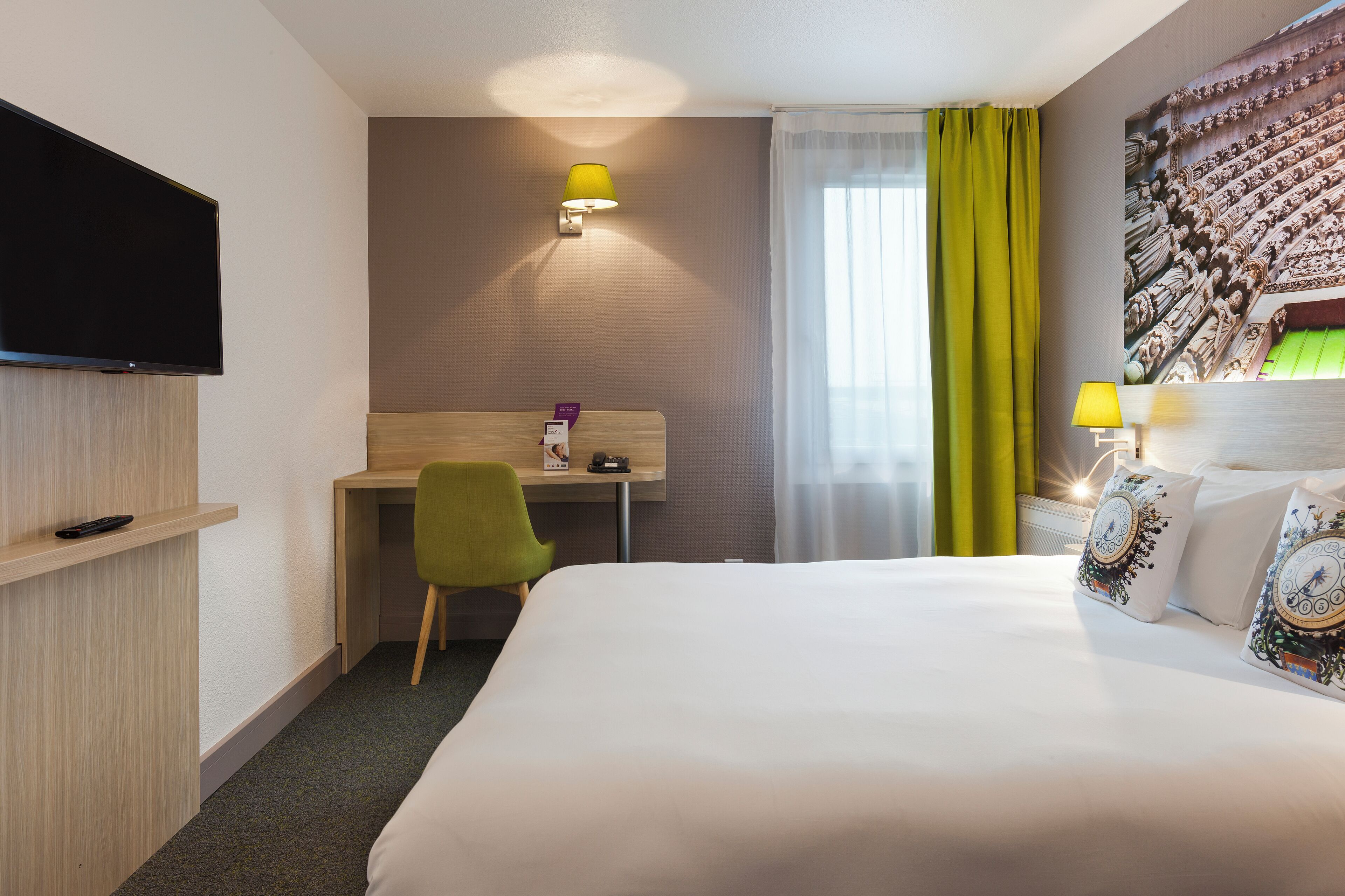 A hotel room with a bed, a desk, a chair, a television, and a wall-mounted lamp.