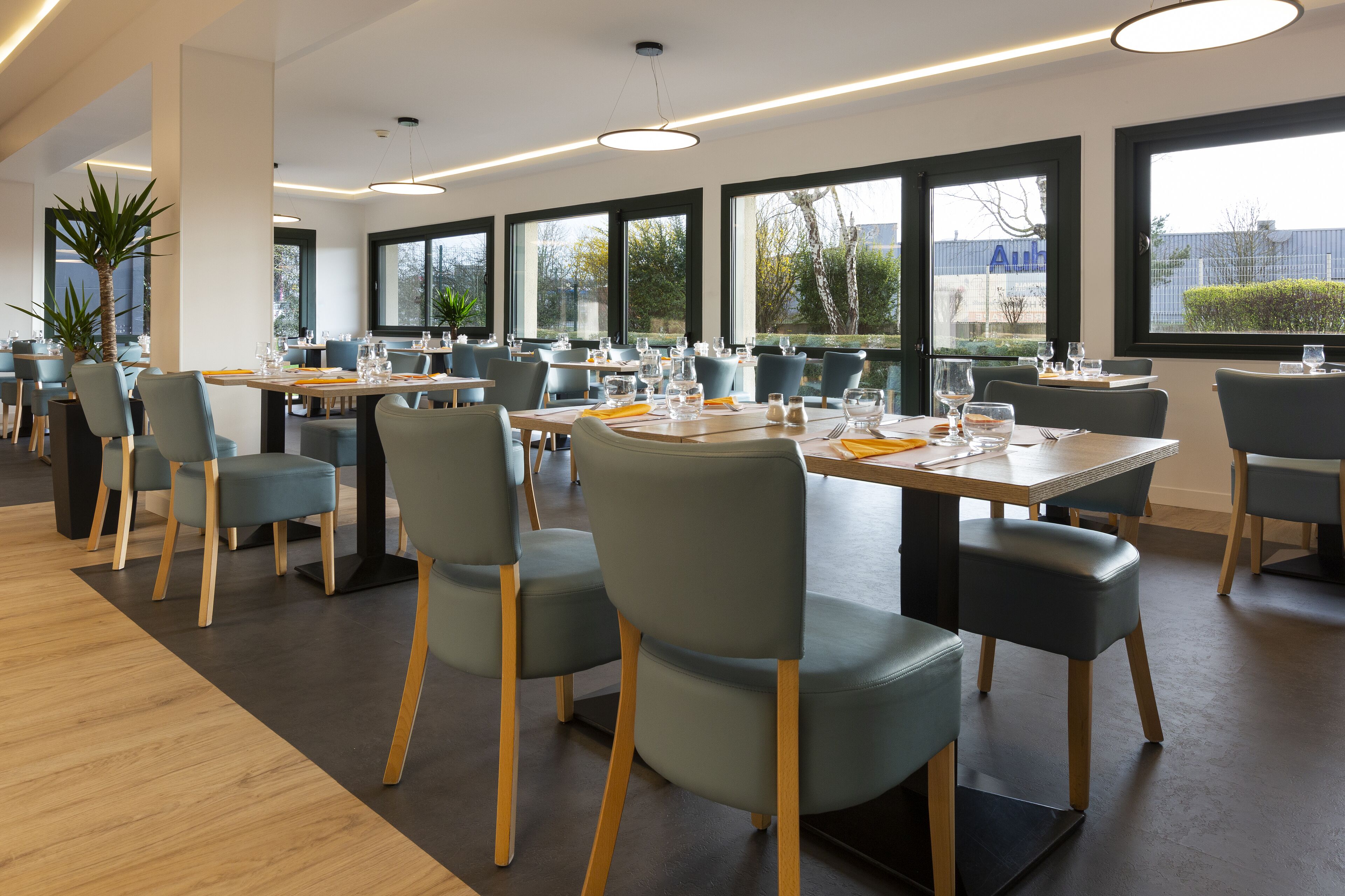 Un espace repas moderne doté de plusieurs tables et chaises, de grandes fenêtres et de plantes d’intérieur.